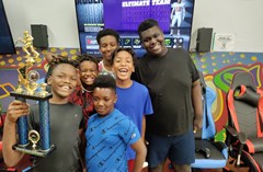 Groups of kids smile in front of TV with gaming chairs in the back ground. One holding a trophy up.