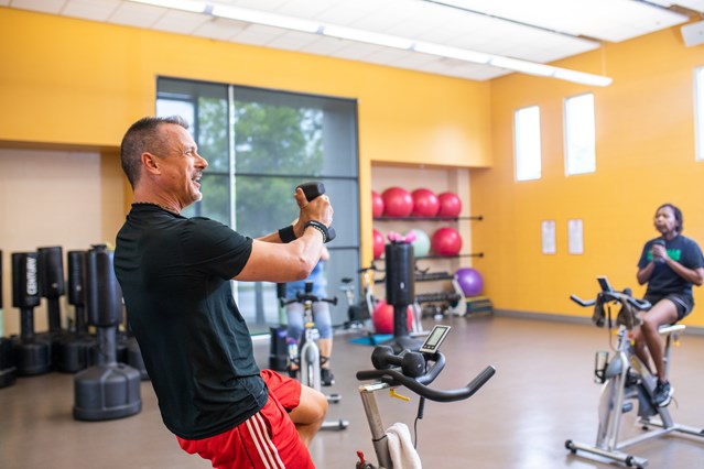 man on Spin bike on right hand side with hand weight in his hands. Woman in background to the right on a spin bike with a weight as well.