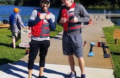 Two adults with lifejackets stand in the foreground with the dock and Lake Crago behind them.