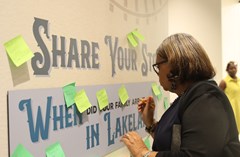 Woman writing on post-it note to add to wall covered in post-it notes and text "Share Your Story. When Did Your Family Arrive in Lakeland?" in Lakeland History & Culture Center exhibit space