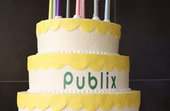 Three-tired white cake with yellow icing scallop decorations, candles on top, and text Publix