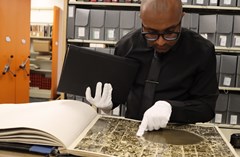 Man wearing white gloves examining large old map book with orange shelving in background