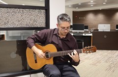 Man playing guitar seated against a wall with computer desks in background
