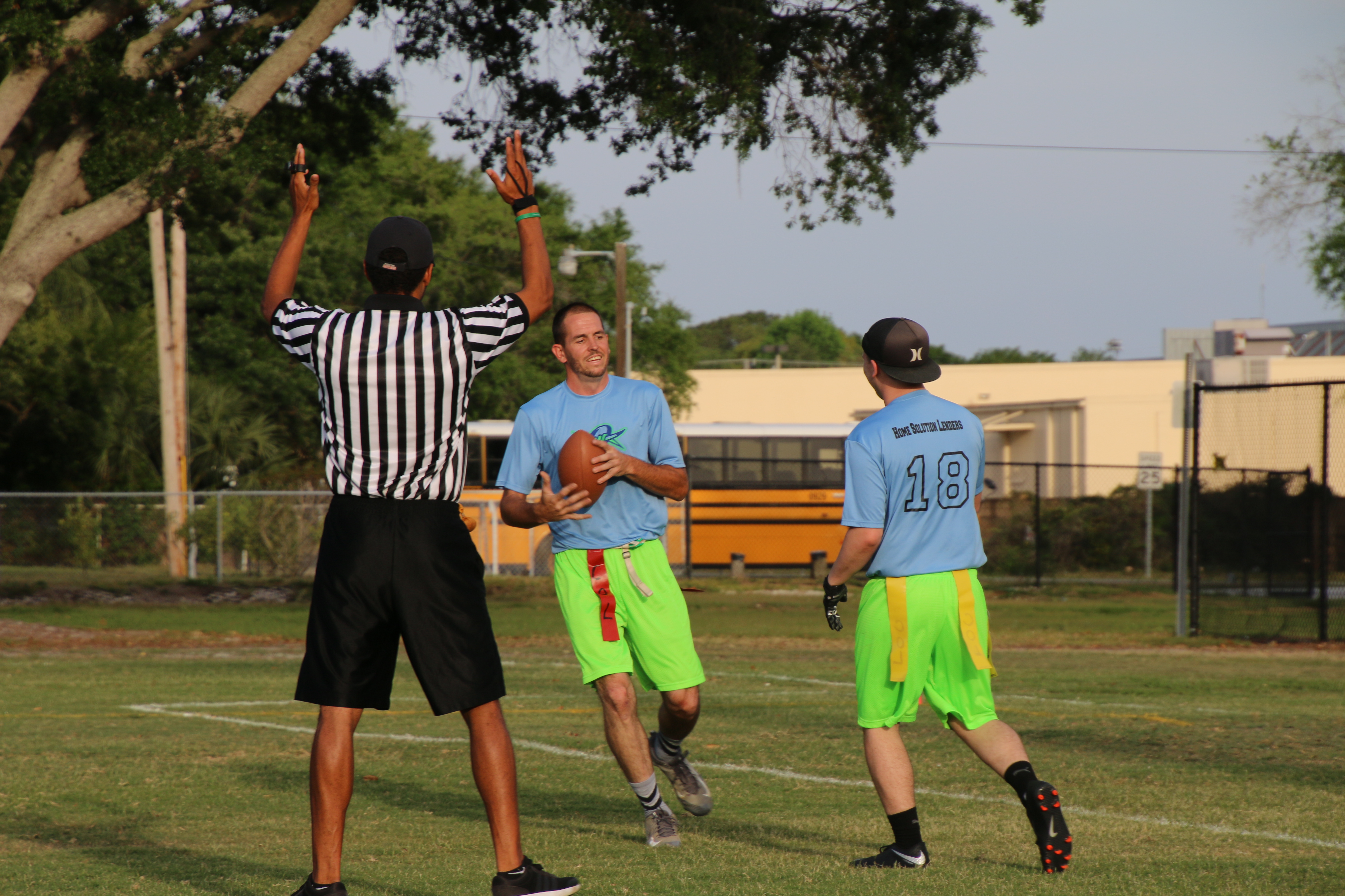 Three men on a field, man in the front with his back turned wearing referee's black and white stripes, other two in the back: One has football in his hand.