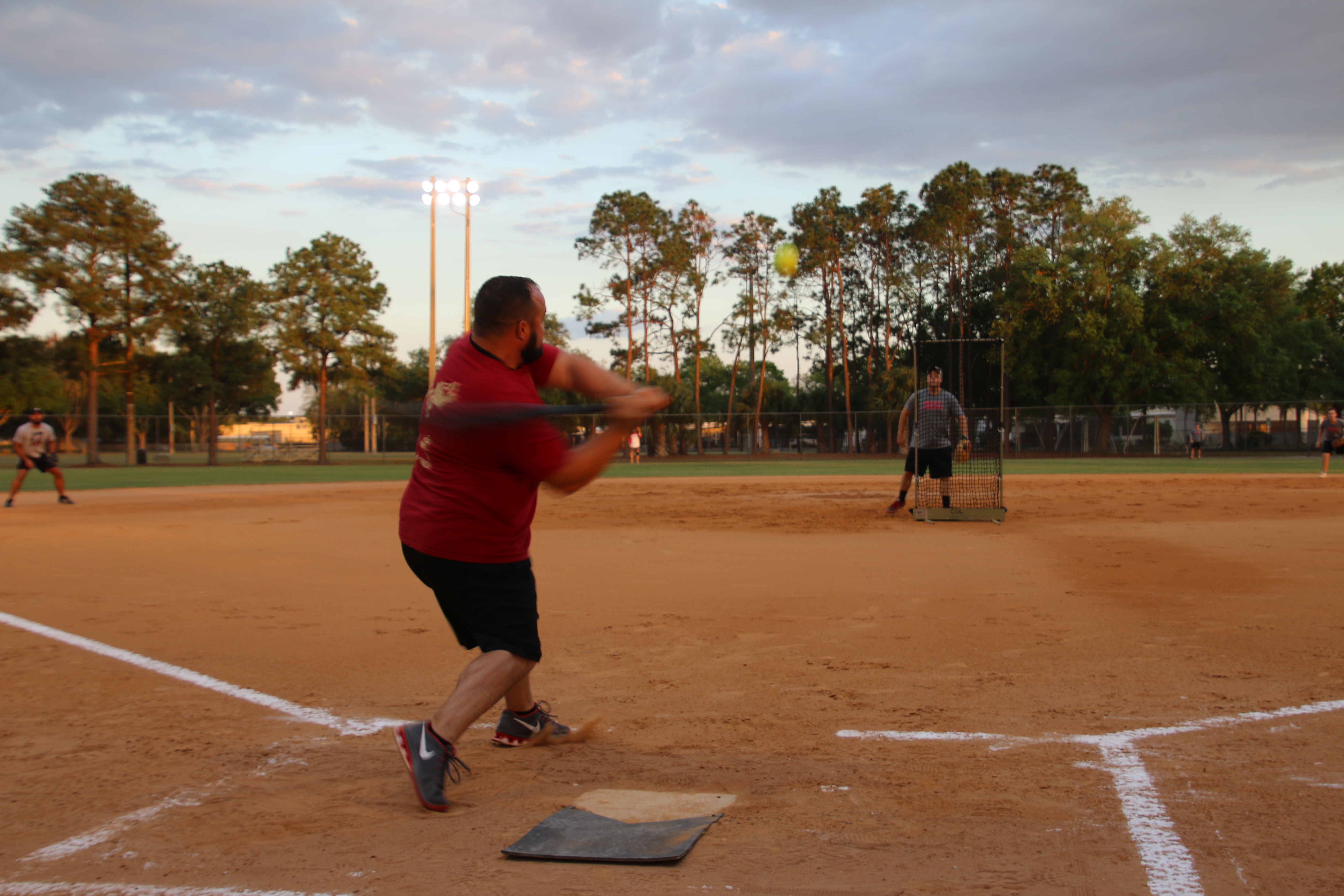 Softball field at Sunset, man in the foreground swinging a baseball bat