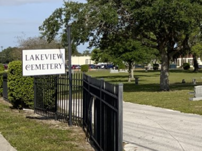 View into Lakeview Cemetery with trees, shrubbery, sign, and wrought iron gate surround; link to Google map to Lakeview Cemetery