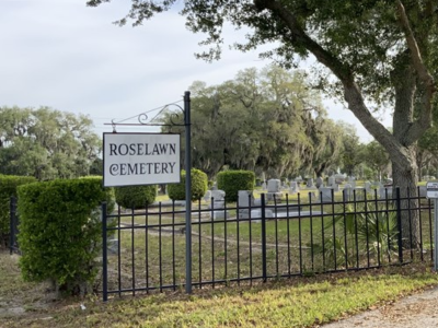 View into Roselawn Cemetery with trees, shubbery, sign, and wrought iron gate surround; link to Google map to Roselawn Cemetery