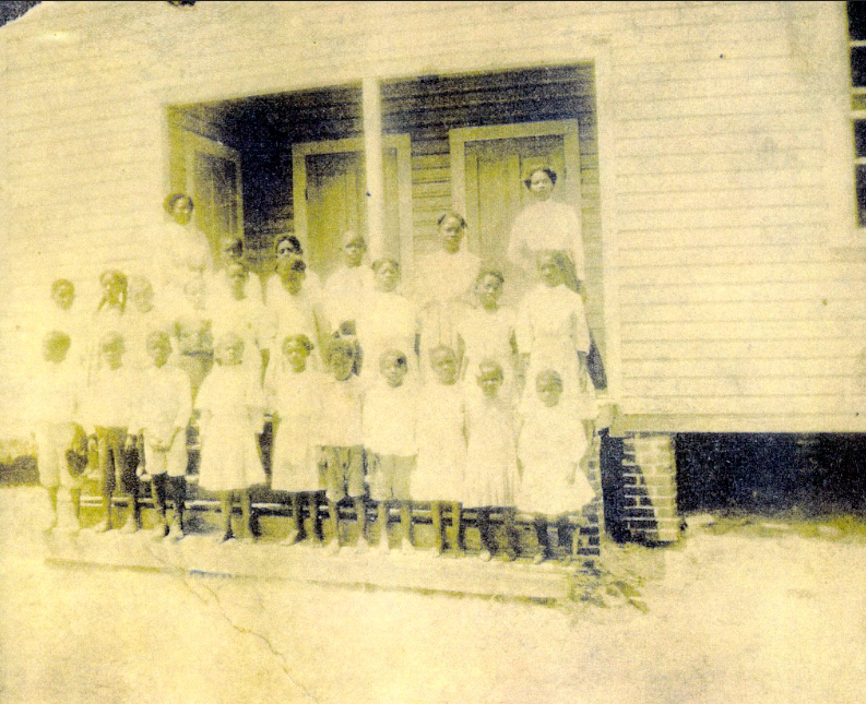 Circa 1930s, students pose outside of the school in the Morehead Community with teacher Mrs. Nettie Adderly on the top right side; link to I Love a Parade album